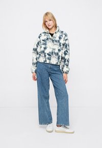 Sudadera de forro polar en patrón de tie-dye blanco y azul marino, con cremallera hasta la mitad en la parte frontal y puños acanalados, combinada con jeans de pierna ancha en azul.