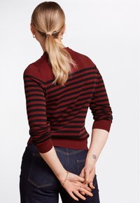 Pull rayé bordeaux et noir avec des poignets côtelés. Le design présente un col rond et une silhouette ajustée. Porté avec un jean sombre.