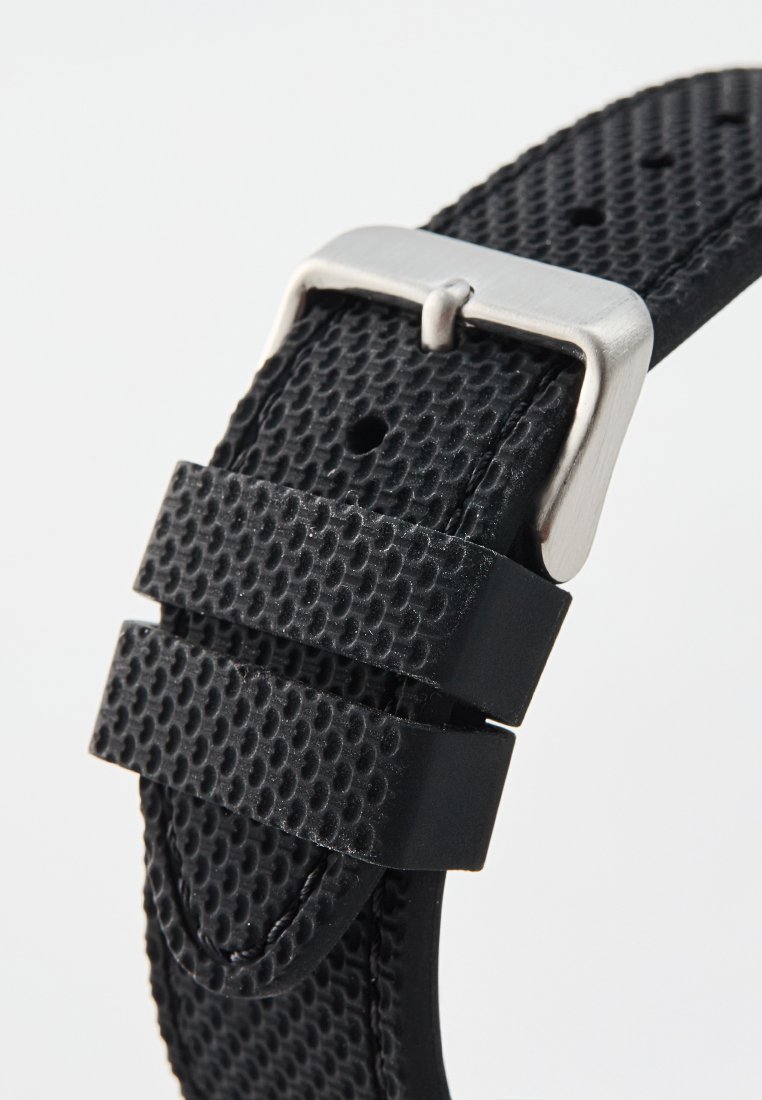 Correa de reloj negra texturizada hecha de goma, con hebilla de metal en tono plateado y dos lazos negros para mantener la correa, con múltiples agujeros de ajuste.