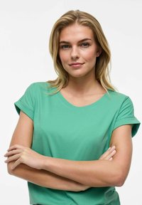 Grön kortärmad t-shirt i mjukt tyg. Har rund halsringning och uppvikta ärmar. Modellen poserar med armarna i kors.