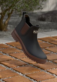 Zwarte waterdichte enkelboot met bruine elastische zijpanelen en een gestructureerde zool, met een logo op de voorkant en een gladde afwerking.