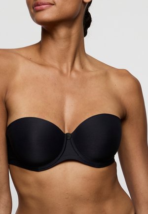 Kobieta w czarnym biustonoszu bez ramiączek, pokazująca górną część tułowia i szyję na jasnym, jednolitym tle.