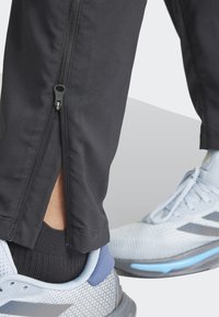 Schwarze Sporthosen mit einem Reißverschluss entlang des Unterschenkels, kombiniert mit hellgrauen Laufschuhen mit Mesh-Textur und blauen Akzenten.