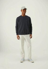 Sweatshirt preto de gola redonda, em mistura de algodão, com punhos e bainha canelados, usado com calças brancas e ténis azul claro, modelo de pé contra um fundo simples.