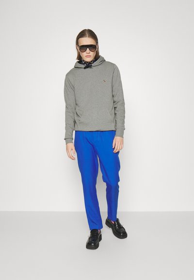 Hanorac gri cu buzunar frontal, pantaloni jogger albaștri și pantofi cu platformă negri. Modelul poartă ochelari de soare supradimensionați și are părul drept.