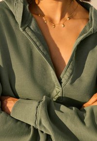Groene hoodie met een V-hals en een voorplacket, voorzien van geribbelde manchetten. Gedragen met een delicate gouden ketting versierd met kleine geometrische hangers.