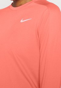 Detailní záběr na korálové sportovní triko s dlouhým rukávem s bílým logem Nike swoosh na hrudi.