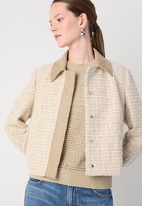 Beige Tweed-Jacke mit einem Wildlederkragen, Druckknöpfen und strukturiertem Finish, kombiniert mit einem hellbeigen Strickpullover und Jeans.