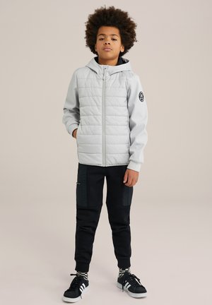 Lys grå polstret jakke med hætte, lynlåslukning og sort logo-patch. Bæres med sorte joggers med sidelommer og sorte sneakers.