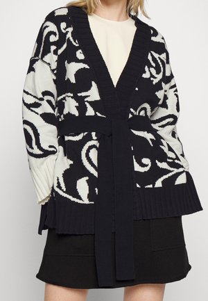 Femme portant un cardigan à motifs noirs et blancs avec une ceinture par-dessus un haut crème et une jupe noire sur un fond uni.
