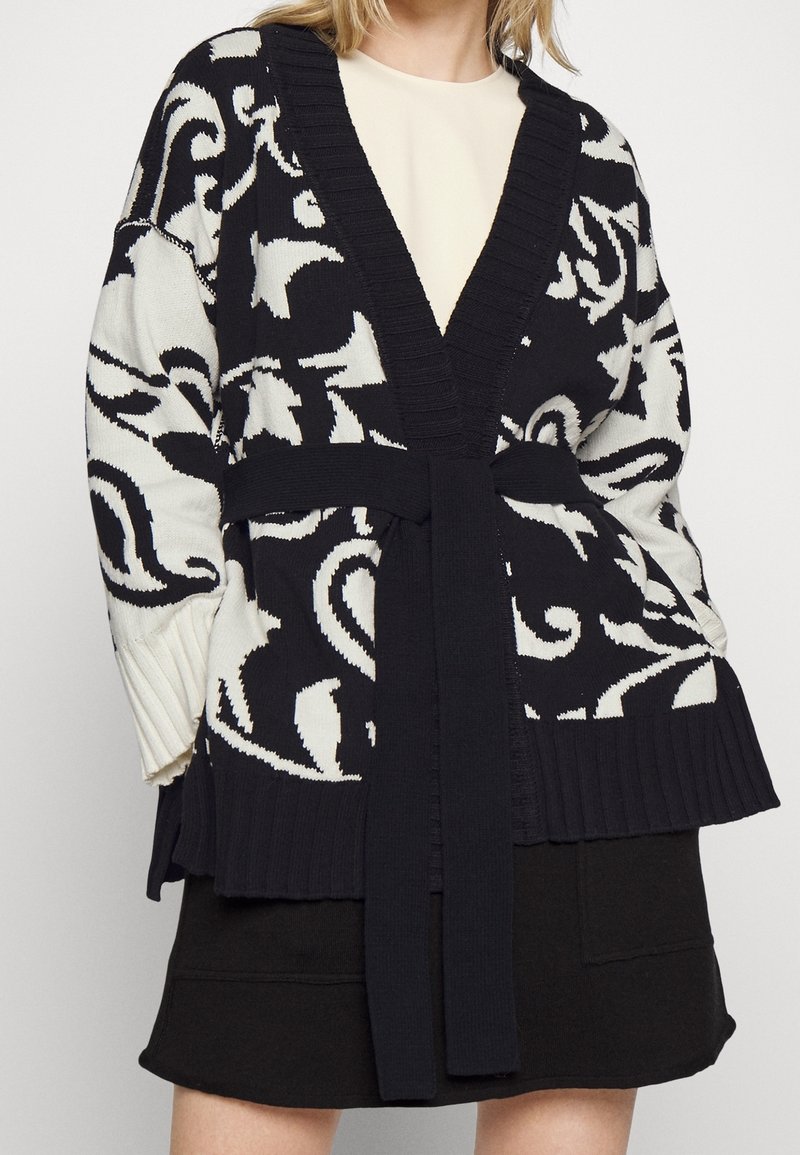 Femme portant un cardigan à motifs noirs et blancs avec une ceinture par-dessus un haut crème et une jupe noire sur un fond uni.