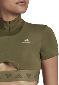 Närbild av en person som bär en olivgrön Adidas kortärmad cropped topp med dragkedja vid kragen och ett märkes-elastiskt band under bysten.