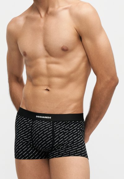 Torso masculin portant un boxer noir à motif DSQUARED2, debout les mains dans le dos sur un fond uni.