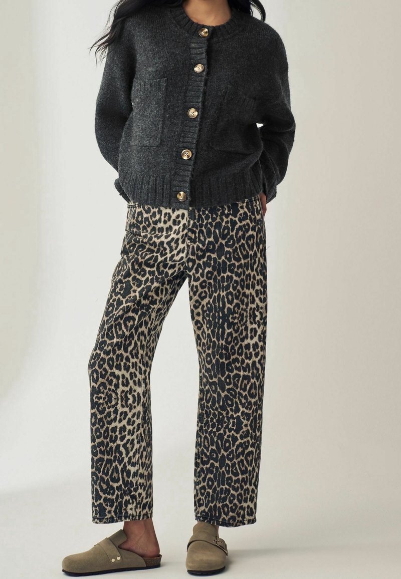 Femme portant un cardigan gris foncé boutonné, un pantalon large imprimé léopard et des chaussures beiges à enfiler avec des boucles, debout les mains derrière le dos.