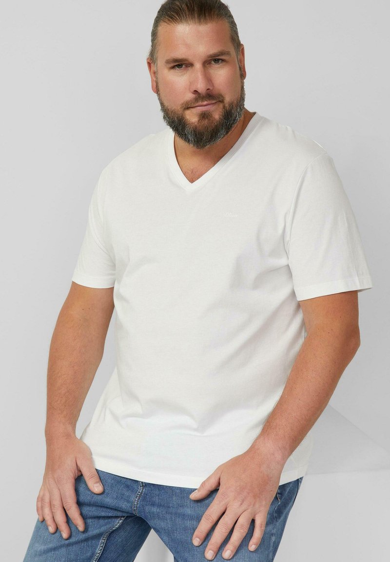 Baardige man met naar achteren gebonden haar, gekleed in een wit T-shirt met V-hals en blauwe jeans, poserend tegen een effen lichtgrijze achtergrond.