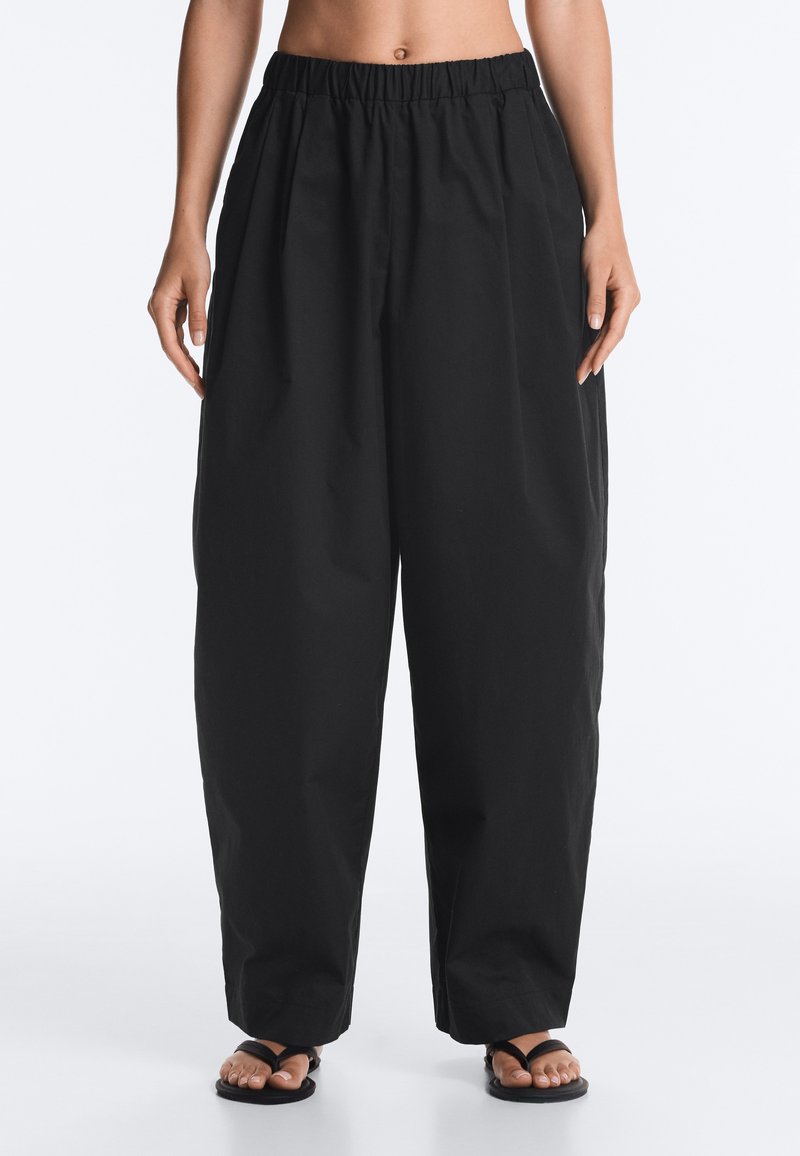Personne portant un pantalon noir large avec une taille élastique et des sandales noires, debout devant un fond blanc uni.