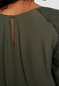 Blusa verde scuro con chiusura a bottone a forma di chiave nella parte superiore della schiena e maniche trasparenti testurizzate con motivi a zigzag.