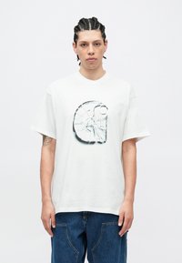 Biele nadrozmerné tričko s grafickou potlačou pripomínajúcou logo rozbitého skla. Krátke rukávy a voľný strih. Neformálne rifľové nohavice.