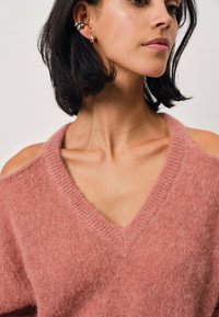 Roze gebreide trui met een V-hals en cold shoulder ontwerp. Zachte textuur, losse pasvorm en geribbelde details bij de halslijn. Zilveren oorbellen zichtbaar.