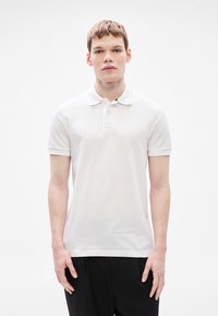 Άσπρο πόλ�ο μπλουζάκι από βαμβάκι με υφασμένη υφή, διαθέτει πατιλέτα με τρία κουμπιά και λεπτομέρεια λογότυπου στο εμπρόσθιο μέρος. Το μοντέλο φοράει μαύρο παντελόνι.