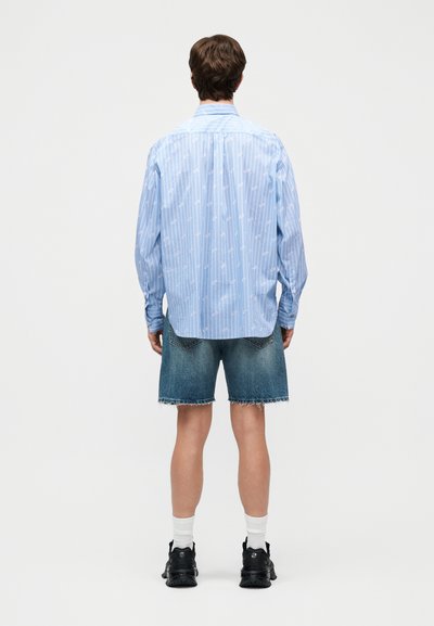 Vīrietis stāv ar muguru redzamu, ģērbies gaiši zilu svītrainu garroku krekli, džinsa šortos, baltās zeķēs un melnās sporta kurpēs.