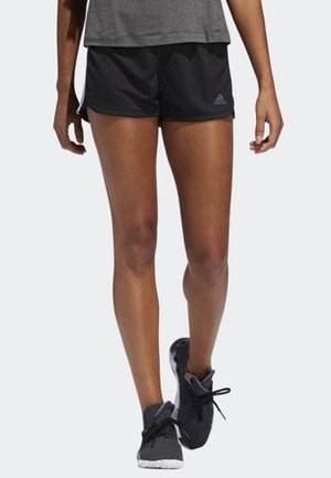 Piernas de una persona que lleva pantalones cortos deportivos negros Adidas, camiseta gris y zapatillas negras de correr caminando hacia adelante sobre un fondo claro.