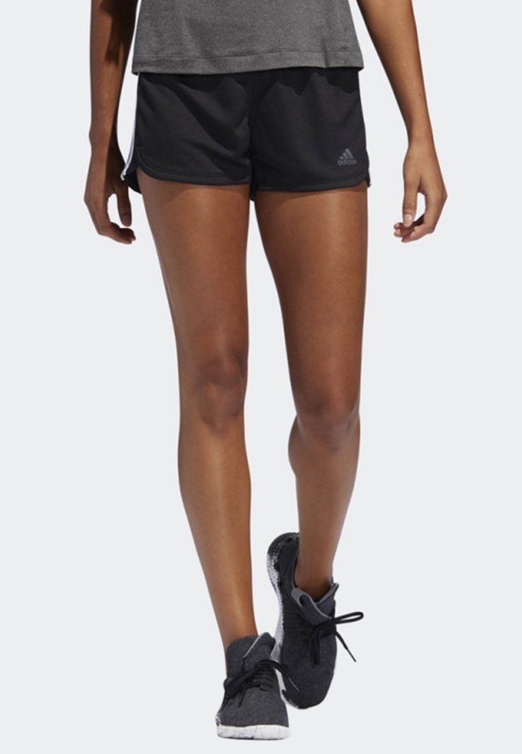 Jambes d'une personne portant un short de sport Adidas noir, un t-shirt gris et des chaussures de course noires marchant vers l'avant sur un fond clair.