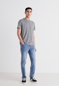 Camiseta gris de manga corta, jeans de mezclilla azul claro y zapatillas marinas con acentos blancos, el modelo se mantiene de manera casual con las manos en los bolsillos.