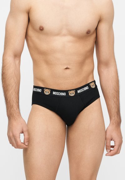 Αρσενικός κορμός που φοράει μαύρα εσώρουχα Moschino με πρόσωπα αρκούδας και το όνομα της μάρκας στη μέση, πάνω σε απλό φωτεινό φόντο.