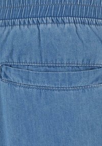 Lyseblåt denimstof med en glat overflade, der har en baglomme syet langs sømmen, fremhævet af synlige sømme.