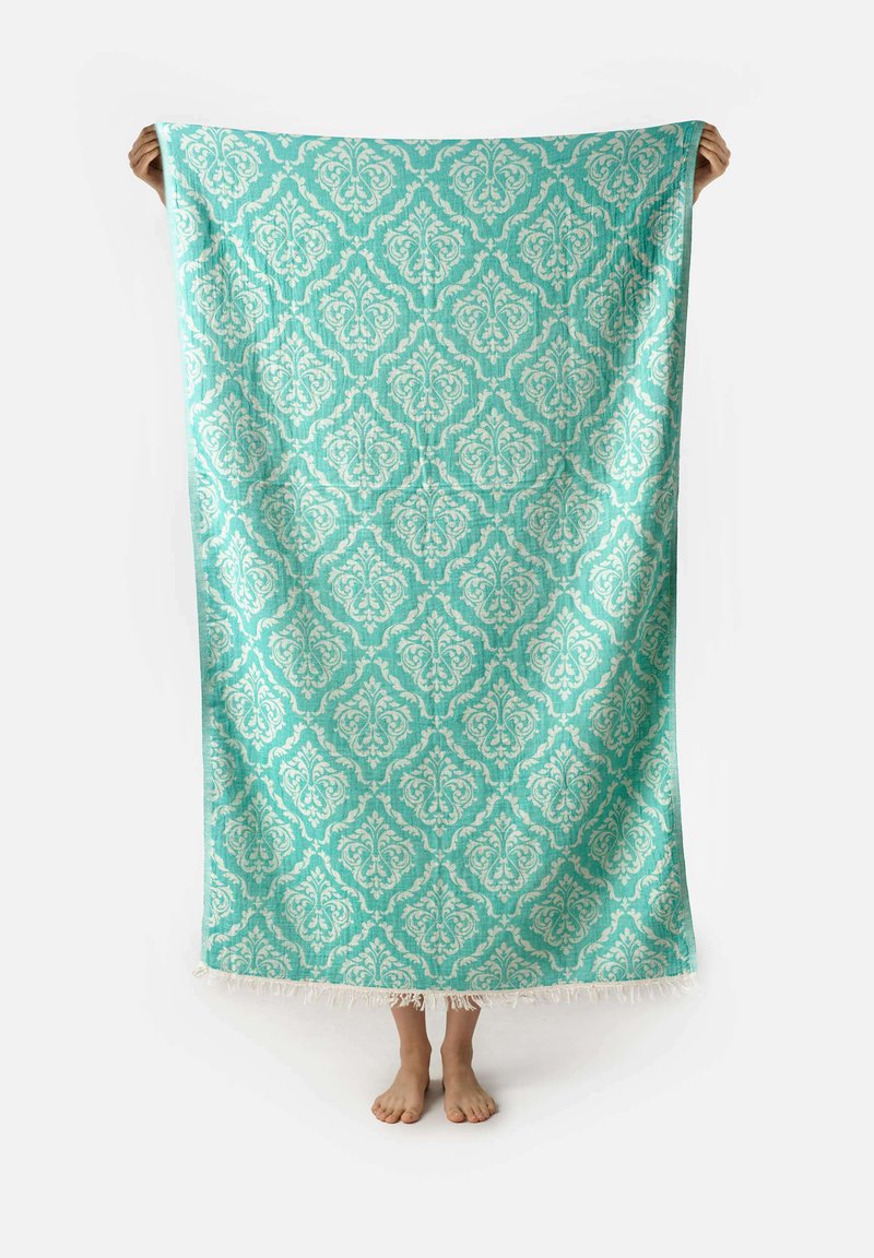 Coperta in turchese con motivi intricati color crema e bordi frangiati, tenuta in alto da una persona. Tessuto leggero, forma rettangolare.