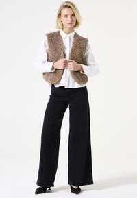 Vrouw in een witte blouse met knopen, een bruine gestructureerde vest, brede zwarte broek en zwarte puntige hakken, staande tegen een witte achtergrond.