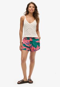 Canottiera bianca all'uncinetto con scollo a V, abbinata a pantaloni corti rosa con motivi di foglie tropicali verdi. La modella indossa sandali scuri.