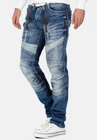 Jeans in denim blu con molte tasche con zip, toppe chiare e scure in contrasto, vestibilità slim e cuciture rinforzate. Accompagnati da sneaker bianche.