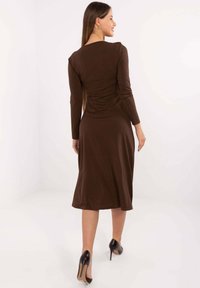 Brązowa sukienka z długim rękawem, o dopasowanym gorsecie i rozkloszowanym dole, z marszczonym tyłem. Noszona z czarnymi szpilkami.