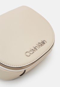 Beżowa torebka na ramię z eko skóry z zaokrągloną klapką, srebrnym logo "Calvin Klein" i kontrastującym przeszyciem. Kompaktowy i usztywniony fason.