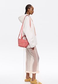 Borsa rosa con design strutturato, superficie testurizzata, manici corti e una lunga tracolla regolabile. Indossata sopra un abito leggero e trasparente.