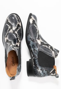 Botas de tobillo negras con un patrón de piel de serpiente en gris, blanco y azul. Cuentan con paneles laterales elásticos y un tacón bajo. Nombre de la marca visible en el interior.