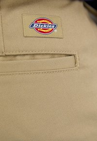 Tissu en twill beige avec une poche cousue, comportant une étiquette rectangulaire avec un design circulaire rouge et jaune et le mot "Dickies".