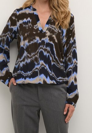 Femme portant un chemisier col en V avec des motifs abstraits bleus, noirs et beiges et un pantalon gris ajusté, main dans la poche, sur fond uni.