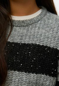 Jasnoszary dziany sweter z grubą czarną taśmą zdobioną cekinami, okrągły dekolt i ściągacze przy rękawach. Teksturowana tkanina z kontrastującymi kolorami.