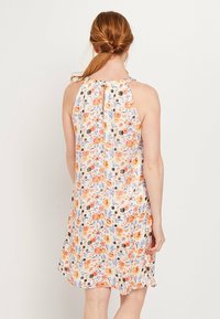 Femme aux cheveux roux avec une queue de cheval tressée, portant une robe sans manches à fleurs, arrivant au genou, ornée de fleurs orange, violettes et noires, de dos.