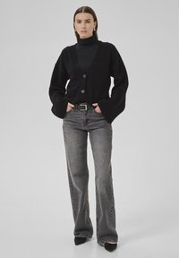 Femeie în picioare, îmbrăcată cu un cardigan negru cu nasturi peste un pulover negru cu guler înalt, blugi gri drepti cu curea și pantofi negri cu vârful ascuțit, pe un fundal simplu.