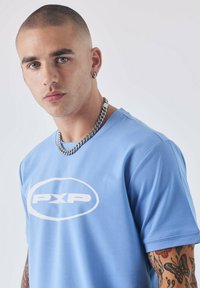 Camiseta de manga corta azul claro con el logo blanco "PXP" en la parte delantera. Diseño de cuello redondo, textura suave y un ligero brillo. Lleva una cadena como collar.