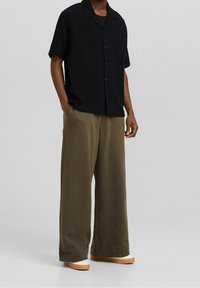 Svart kortärmad skjorta med knappar och texturerad tyg, tillsammans med lösa olivgröna byxor och ljusa skor.