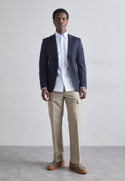 Marineblazer, lyseblå skjorte med knapper, beige cargo-bukser. Brune sko med tekstureret overflade. Minimalistisk design, skræddersyet pasform.