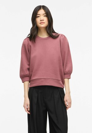 Młoda kobieta z długimi ciemnymi włosami, ubraną w mauve bluzkę z bufkami i czarne plisowane szerokie spodnie, stojąca na białym tle.