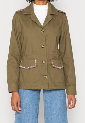 Kurtka w kolorze oliwkowej zieleni zapinana na guziki z kwiatową lamówką na pagonach i klapach kieszeni, noszona z jasnoniebieskimi jeansami.