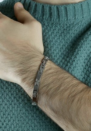 Bracciale in metallo argentato con sezioni rettangolari collegate, appoggiato su un polso con capelli castano chiaro, su uno sfondo di un maglione verde texture.