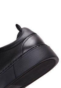 Tomaia in pelle nera di una sneaker con tacco, suola in gomma testurizzata e linguetta in tessuto, in primo piano su sfondo bianco.
